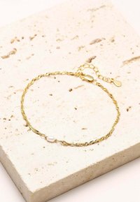 Goldener, gedrehter Armreif mit einer einzelnen Perlenakzentuierung und einem Verschluss mit einer verstellbaren Kette. Vor einem strukturierten Stein-Hintergrund drapiert.