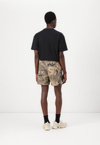 Schwarzes T-Shirt mit kurzen Ärmeln kombiniert mit beigen Shorts, die ein schwarzes Blumenmuster aufweisen. Das Modell trägt schwarze Socken und hellbeige Sneakers.