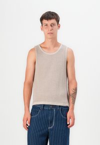 Top sin mangas de color beige con textura acanalada y amplios agujeros para los brazos, combinado con jeans azul a rayas. El top presenta un cuello redondo y una tela texturizada.