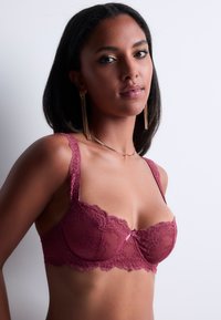 Soutien-gorge en dentelle bourgogne, avec des bords festonnés, un nœud en satin au centre et des bretelles réglables. La texture translucide enrichit le design.