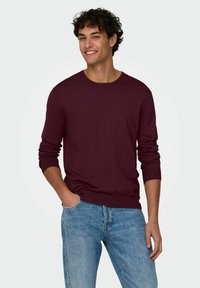 Maglione a maglia bordeaux con maniche lunghe, scollatura rotonda e polsini a coste, abbinato a jeans blu chiaro per un look casual.