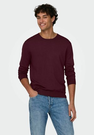 Maglione a maglia bordeaux con maniche lunghe, scollatura rotonda e polsini a coste, abbinato a jeans blu chiaro per un look casual.