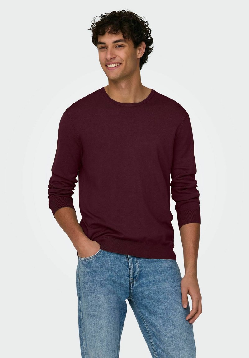 Maglione a maglia bordeaux con maniche lunghe, scollatura rotonda e polsini a coste, abbinato a jeans blu chiaro per un look casual.