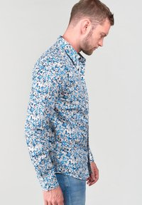 Camicia abbottonata con un motivo floreale blu e bianco, realizzata in tessuto leggero, con colletto e maniche lunghe, mostrata di lato.