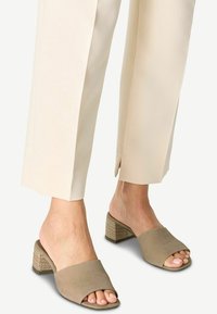 Mules beiges à bout ouvert avec une texture en daim doux, présentant un petit talon carré en matériau tissé et un design de bride supérieure incurvée.