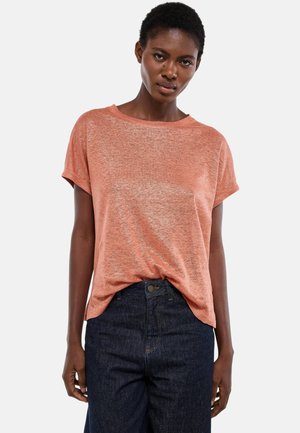 Femme portant un haut ample à manches courtes de couleur saumon légèrement rentrée dans un jean taille haute bleu foncé, debout devant un fond uni.