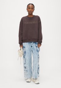 Tumši brūns džemperis ar uzrakstu "DOLCE FAR NIENTE". Gaiši zilas darba stila džinsa bikses ar vairākām kabatām. Baltas sporta kurpes un neliela bēša somiņa.