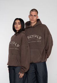 Hnedé oversized mikiny s kapucňou a predným vreckom, s grafickým dizajnom nápisom "MULTIPLY" a rokom "2015." Spárované s tmavými nohavicami.