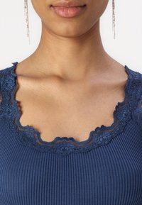 Navyblauw geribd bodysuit met een gegolfde kantenkraag. De kant heeft bloemenpatronen en een zachte textuur, wat het ontwerp van het kledingstuk versterkt.