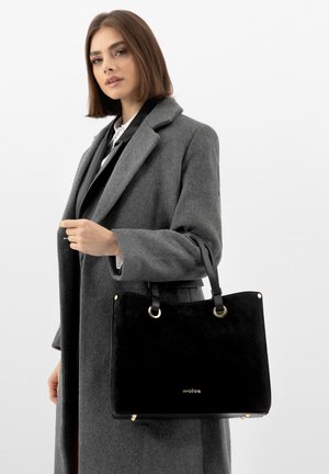 Vrouw in een grijze jas die een zwarte boodschappentas met gouden accenten vasthoudt tegen een effen witte achtergrond.