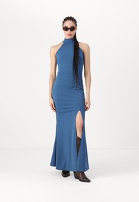 Rochie fără mâneci, de culoare albastră, cu guler înalt, fanta pe lateral și o siluetă ajustată. Materialul este neted, cu un finisaj mat. Asortată cu botine maro cu toc.