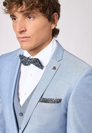 Lichtblauw pak met een gestructureerde afwerking, wit shirt en een donker bloemenstrik. Voorzien van een pattenzakdoek en een logo op het revers.