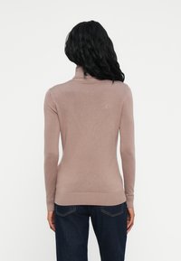 Donna con lunghi capelli neri che indossa un maglione a collo alto color malva e jeans blu scuro, vista di spalle su uno sfondo bianco.