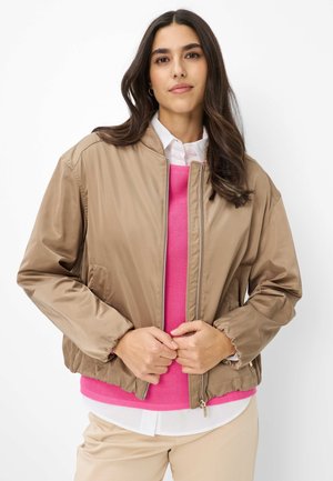 Frau mit langen, dunklen Haaren trägt eine beige Bomberjacke über einem leuchtend pinken Pullover und einem weißen Hemd, posiert mit Händen in der Nähe des Reißverschlusses der Jacke.