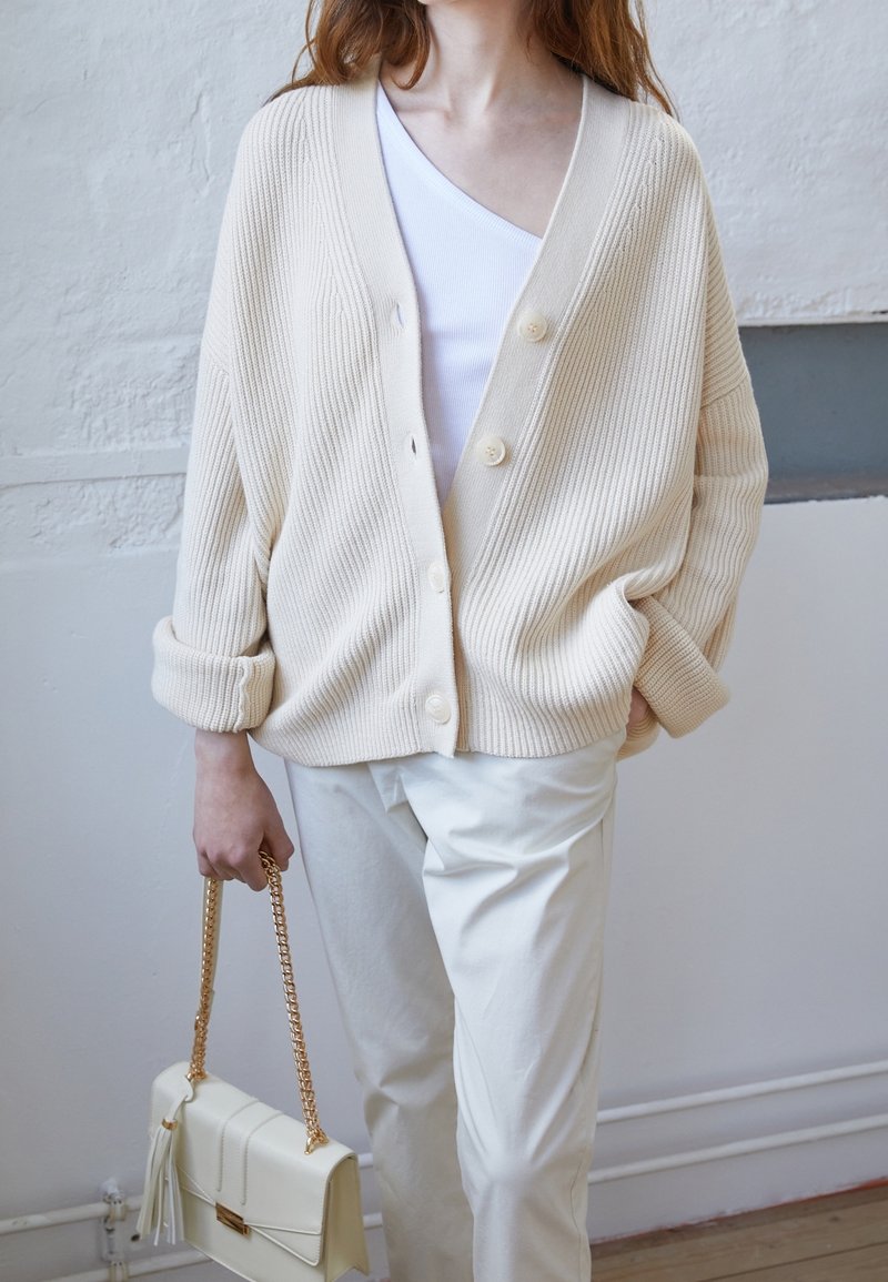 Femme portant un cardigan côtelé crème par-dessus un haut blanc et un pantalon blanc, tenant un sac à main crème avec une bandoulière chaîne dorée.
