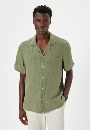 Camisa verde de manga corta hecha de tejido texturizado, con un cierre de botones en la parte delantera, cuello y un bolsillo de parche en el lado izquierdo del pecho.