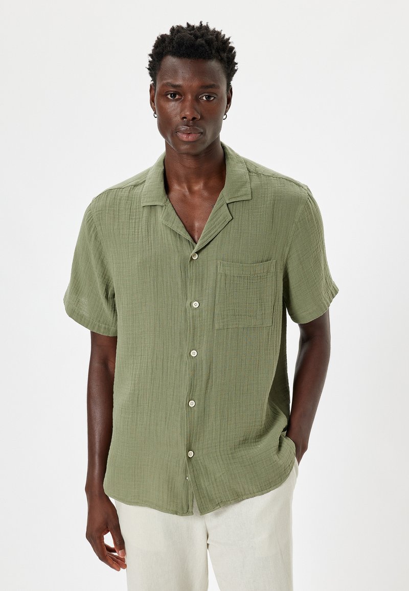 Camisa verde de manga corta hecha de tejido texturizado, con un cierre de botones en la parte delantera, cuello y un bolsillo de parche en el lado izquierdo del pecho.