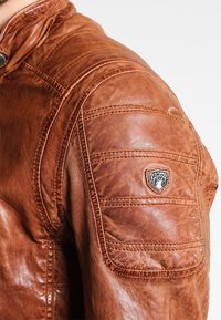 Giacca di pelle marrone con design delle spalle trapuntato, finitura testurizzata e una patch con dettagli del logo sul maniche sinistra.