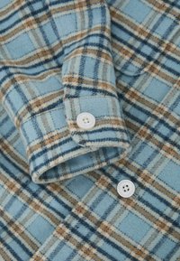 Manche et poignet de chemise en flanelle bleue avec boutons blancs, présentant un motif à carreaux beige et bleu marine.