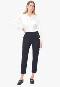 Pantalons taille haute marine avec quatre boutons dorés sur le côté, associés à un blouse blanche à manches longues et des mocassins noirs.