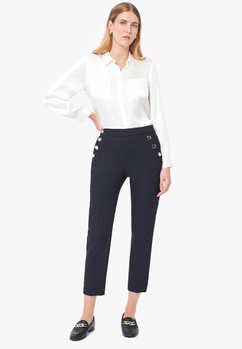 Pantalons taille haute marine avec quatre boutons dorés sur le côté, associés à un blouse blanche à manches longues et des mocassins noirs.