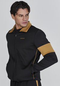 Veste zippée noire avec un col et des accents de manches brun doré. Elle présente un logo sur la poitrine et une coupe ajustée. Texture de tissu lisse.