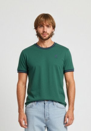 Mann mit hellbraunen Haaren, der ein dunkelgrünes kurzärmliges T-Shirt mit marineblauen Details und hellblaue Jeans trägt, steht vor einem schlichten weißen Hintergrund.