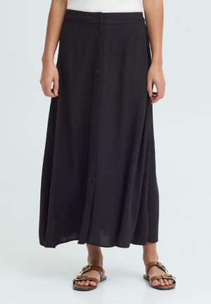 Kvinne iført et svart midi-skjørt med knapper foran, kombinert med brune sandaler med spenne, vist fra livet til føttene mot en ensfarget bakgrunn.