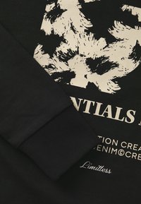 Black sweatshirt featuring a textured, ribbed cuff and a large cream graphic with abstract motifs. Branding text is visible below the design.