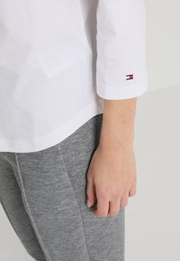Camicia bianca a maniche lunghe con dettagli ai polsini, abbinata a pantaloni sportivi grigi. Include un piccolo logo multicolore sulla manica della camicia.