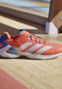 Scarpa da corsa arancione e blu con strisce e lacci bianchi, appoggiata su un pavimento in legno chiaro con sfondo sfocato.