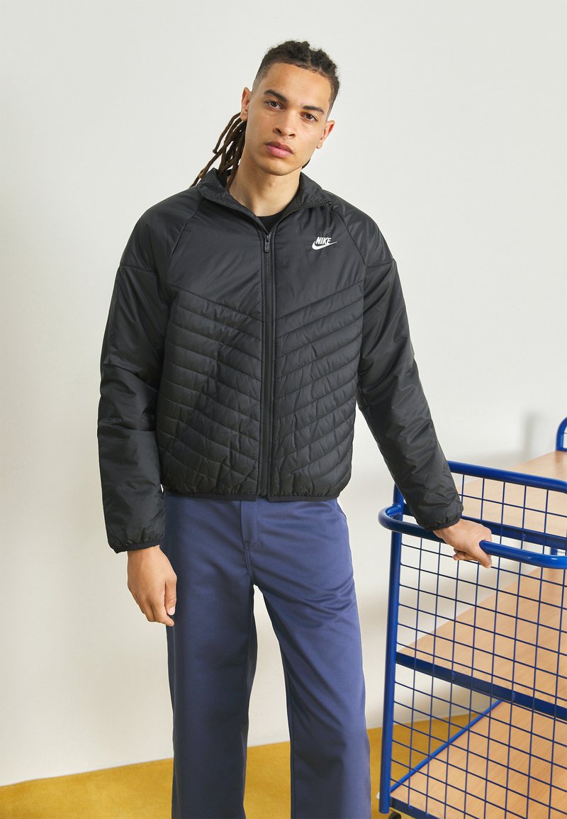 Giacca a vento nera trapuntata con zip frontale, piccolo logo Nike sul petto e polsini elastici. Indossata sopra pantaloni blu, su uno sfondo neutro.