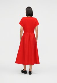 Κόκκινο βαμβακερό φόρεμα με κοντά μανίκια, στρογγυλό ντεκολτέ και ζωνάκι στη μέση, που ρέει σε φούστα μήκους έως τη μέση της γάμπας· φοριέται με μαύρα παπούτσια.