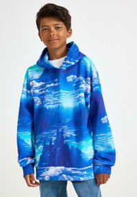 Dreng iført en blå hættetrøje med et mønster af skyer og bylandskab, parret med lyseblå jeans, der står foran en ensfarvet hvid baggrund.