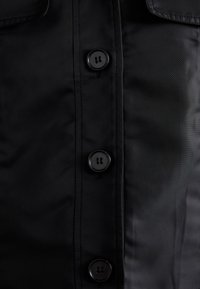 Zwarte button-up overhemd gemaakt van gladde stof met vier zwarte plastic knopen aan de voorkant en een gestructureerde kraag.