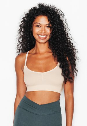Femme aux longs cheveux bouclés portant un crop top beige et des leggings taille haute vert foncé, souriante sur un fond blanc.