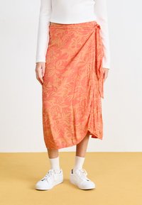 Rosa omslagskjørt med et tropisk bladmønster. Laget av lett stoff, med en sidetilknyting og en buet nederkant. Kombinert med hvite joggesko.