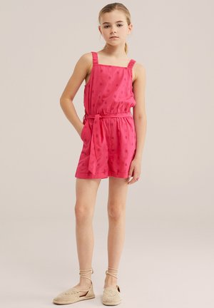 Junges Mädchen steht mit der Hand in der Tasche, trägt einen ärmellosen rosa gepunkteten Overall und beige gewebte Schuhe mit Knöchelband vor neutralem Hintergrund.