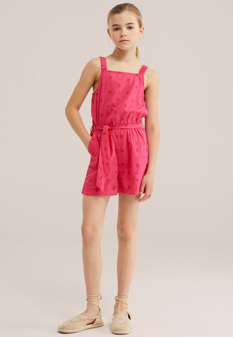 Jeune fille debout, main dans la poche, portant une combinaison sans manches rose à pois et des chaussures beiges tissées avec lacets à la cheville sur fond neutre.