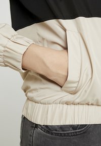 Jasno beżowa i czarna kurtka z elastycznymi mankietami i dołem, z boczną kieszenią po lewej stronie. Gładka, lekko błyszcząca faktura materiału.