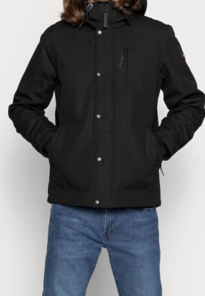 Person iført sort lynlåslukket hættetrøje med trykknapper og forlommer, kombineret med blå jeans.