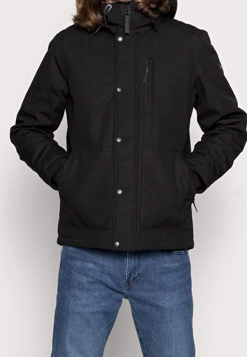 Personne portant une veste à capuche noire zippée avec boutons-pression et poches frontales, associée à un jean bleu.