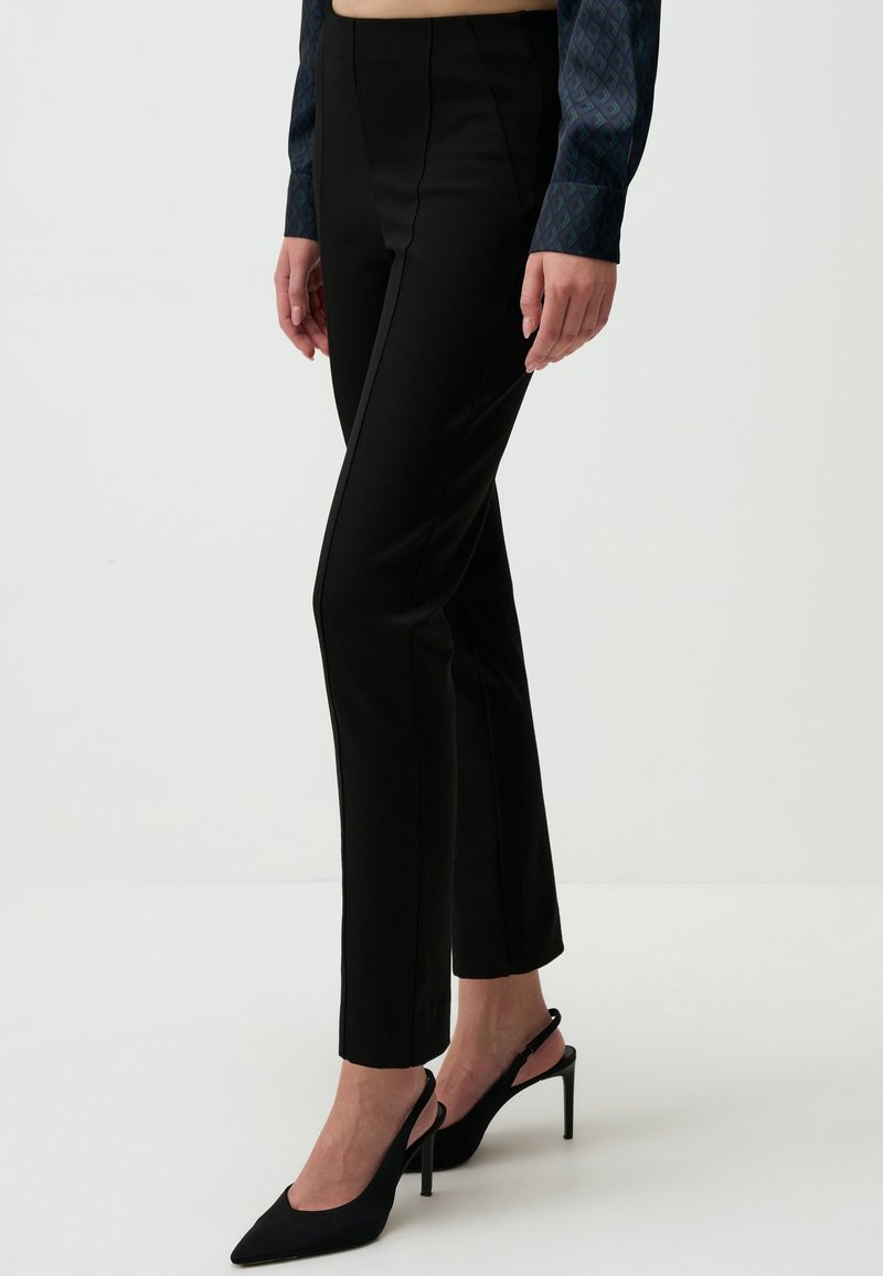 Femme portant un pantalon noir slim et des chaussures slingback noires à talons hauts, debout devant un fond blanc uni.