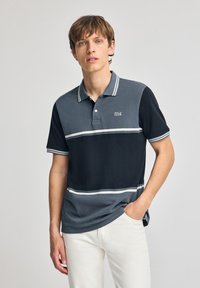 Grå och marinrandig pikétröja med vita accenter, med en treknappslåsning och logotyp på bröstet. Tillverkad av bomullsblandning.