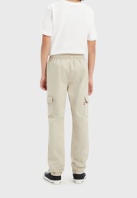 Dziecko ubrane w beżowe spodnie cargo, białą koszulkę i czarne sneakersy, stojące tyłem na jednolitym tle.