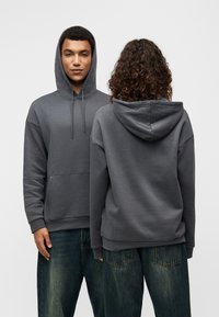 Γκρι φούτερ με κουκούλα, μπροστινή τσέπη, κορδόνι στην κουκούλα και ριμπ μανσέτες. Το υλικό φαίνεται μαλακό με άνετη εφαρμογή, συνδυασμένο με φαρδιά τζιν παντελόνια.