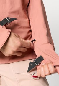 Casaco rosa claro com ferragens pretas e tiras ajustáveis nos punhos. O tecido é suave e ligeiramente texturizado, apresentando um design moderno.