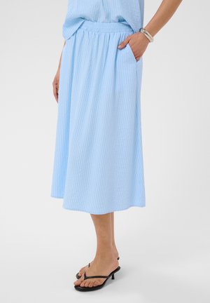 Femme portant une jupe midi bleu clair avec une taille élastique et la main dans la poche, associée à des sandales noires à talons et un bracelet au poignet gauche.