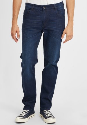 Mørkeblå denimjeans med slim fit, med knappelukking, fem lommer og subtil tekstur. Kombinert med svarte og hvite joggesko.