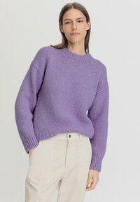 Violetti neulepusero, jossa on pyöreä kaula-aukko, löysä malli ja resorit hihansuissa. Yhdistetty beigen värisiin housuihin, joissa on sivutaskut ja nappi.